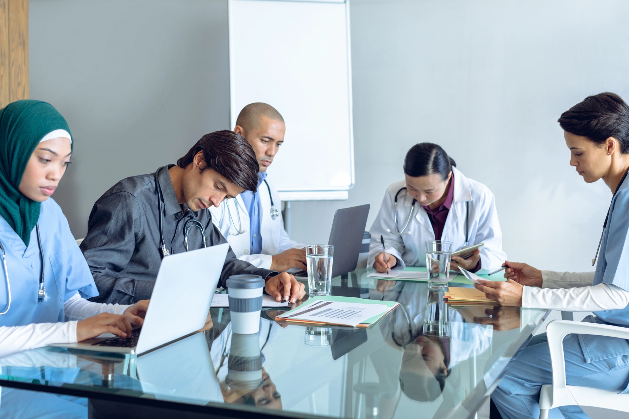 Front view of diverse medical team working together at table in hospital. Coffee cup, glass of water, clipboard, medical folders, and laptop are on the table.