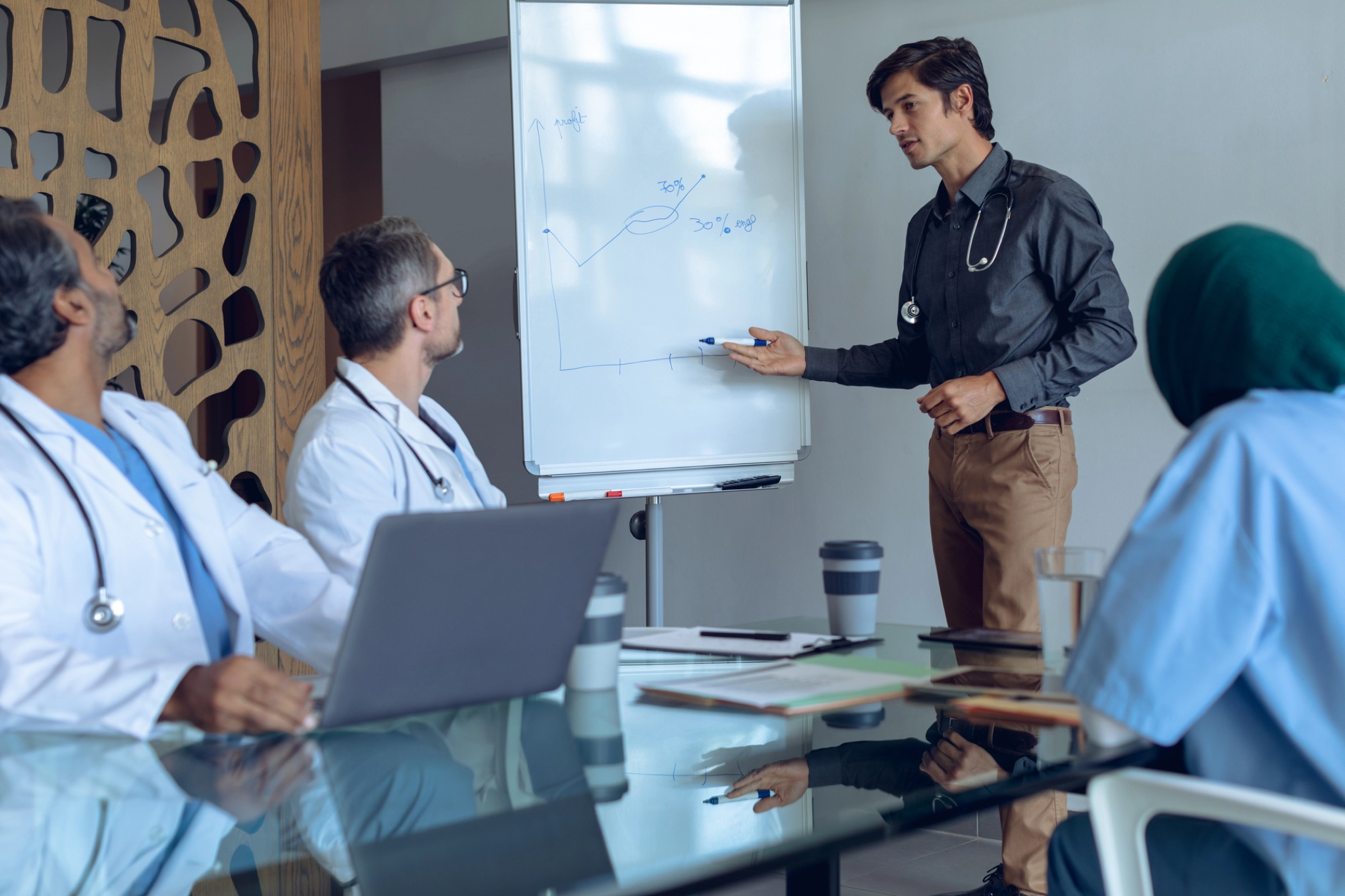 Front view of Caucasian male doctor explaining graph on flip chart in meeting with diverse medical team at hospital. Coffee cup, medical folders, clipboard, digital tablet and laptop are on the table.