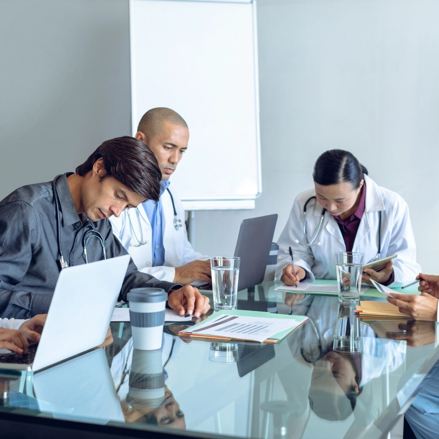 Front view of diverse medical team working together at table in hospital. Coffee cup, glass of water, clipboard, medical folders, and laptop are on the table.